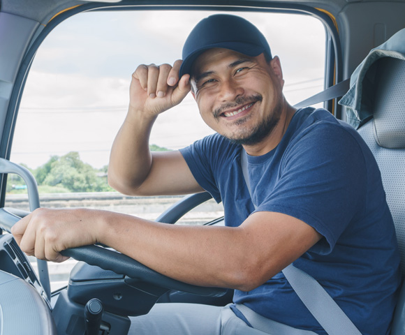 Homem pardo de camiseta e boné azul sentado ao volante do caminhão