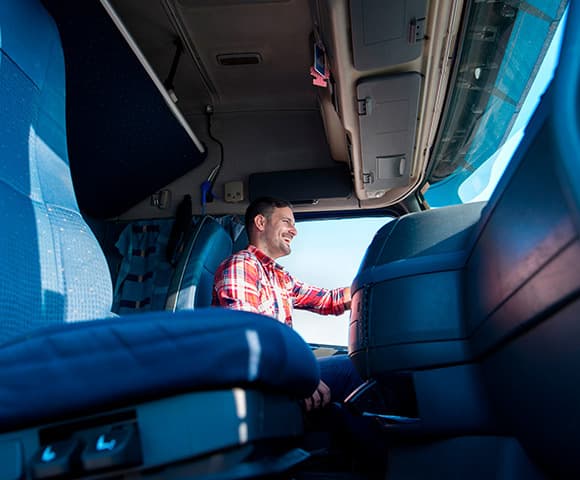 Vemos uma cabine de caminhão com assentos na cor azul painel cor preto e teto cor cinza com um homem sorridente na direção vestindo uma camisa quadriculada vermelha com branca olhando para seu destino