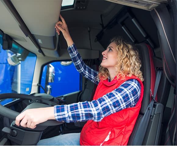 Vemos uma mulher caminhoneira de cabelos ondulados loiros dentro da cabine de caminhão com um mão no volante e a outra apertando algum botão