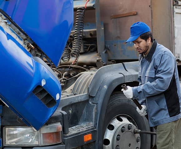 Vemos um homem uniformizado usando luvas com uma ferramenta nas mãos ao lado de um caminhão
